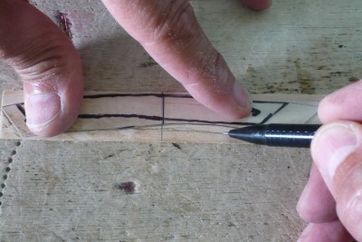 Para los banzos utilizo madera de pino de baja calidad, de la que no tiene prácticamente veta, regruesada a 1 mm de espesor. <br />Con la plantilla, a la que le he marcado una flecha indicando la proa, -se ve debajo de mi dedo índice- marco sobre la madera el contorno del banzo inferior y como siempre, le doy un sobredimensionado, excepto a la línea de unión de banzos que sí respeto escrupulosamente junto con la de la cuaderna maestra.